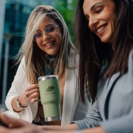 Two smiling women. One is holding a large green mug with a healthcare logo.