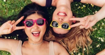 Two smiling people wearing retro neon sunglasses. They are making peace signs and lying on their backs in the grass.