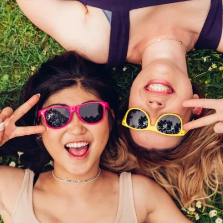 Two smiling people wearing retro neon sunglasses. They are making peace signs and lying on their backs in the grass.