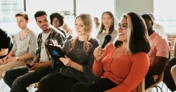 Person holding a microphone while sitting in a meeting with a group of people.  