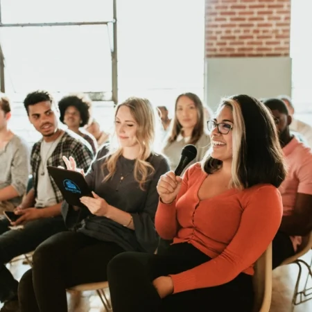 Person holding a microphone while sitting in a meeting with a group of people.  