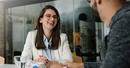 Two people are sitting at a table in an office, engaged in conversation.