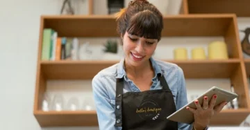 Smiling boutique owner is wearing an apron and holding a tablet. 