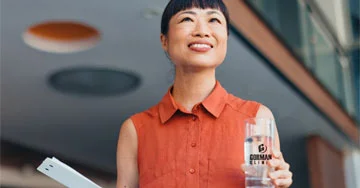 a person in a red dress holding a water bottle and a document, standing in a modern, well-lit office environment.