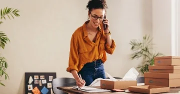 Business owner on the phone. Stacked boxes are on a table next to them. 